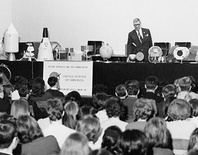 A spacemobile lecturer stands behind a table full of space age models, speaking to an audience of schoolchildren sitting on a gymnasium floor in the 1960s.